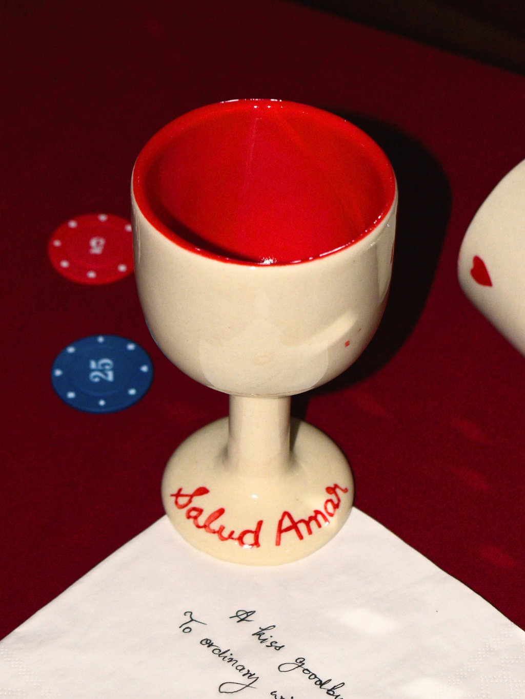 A ceramic wine glass with a cream-colored exterior and a red interior, featuring the text 'Salud Amor' written on the base. It is displayed on a dark red felt surface next to a white napkin with a red lip print and a handwritten message.